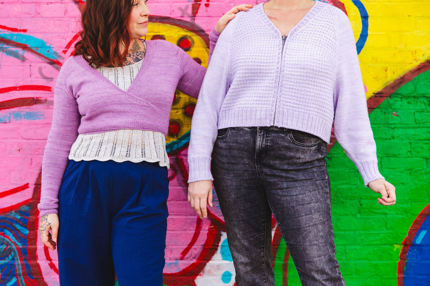 Bess stands, one hand on Jen's shoulder, in front of a wall mural. She wears a purple-pink knit ballet wrap over a lace knit cream tank with navy blue pants. Jen, standing next to her, wears a light purple zip-up sweater, knit with a V-neckline, with a pair of dark acid wash jeans.