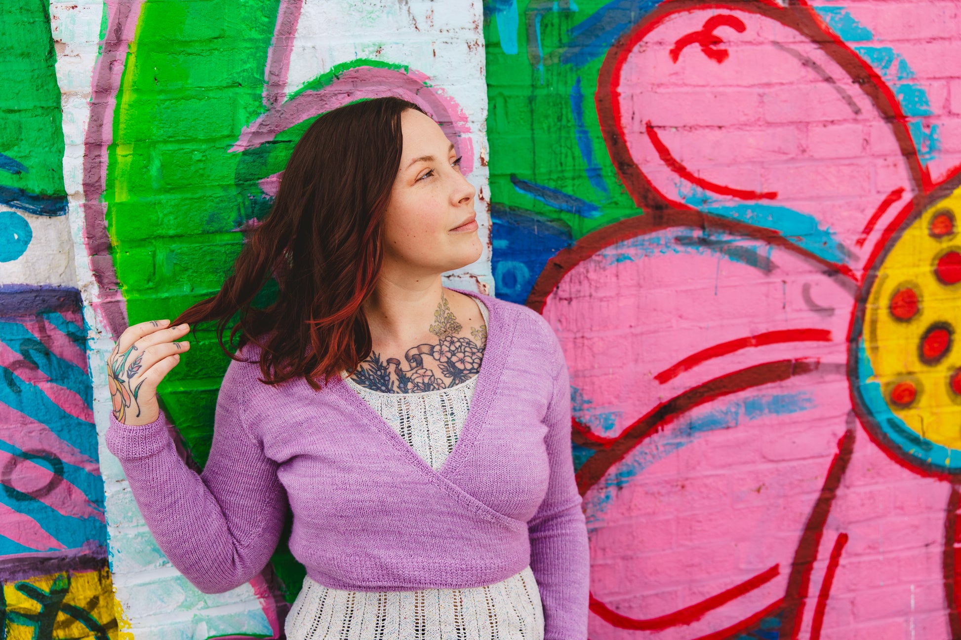 Bess stands in front of a wall mural, one hand playing with her hair. She wears a creamy lace knit tank under a ballet wrap sweater knit with purple-pink yarn.