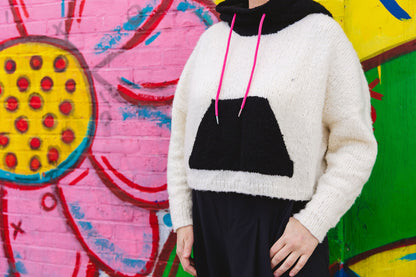 Seen from the shoulders down, Jen wears a white hoodie, knit with a black pouch pocket and hood in a graphic style, with a black skirt. A wall mural can be seen in the background.