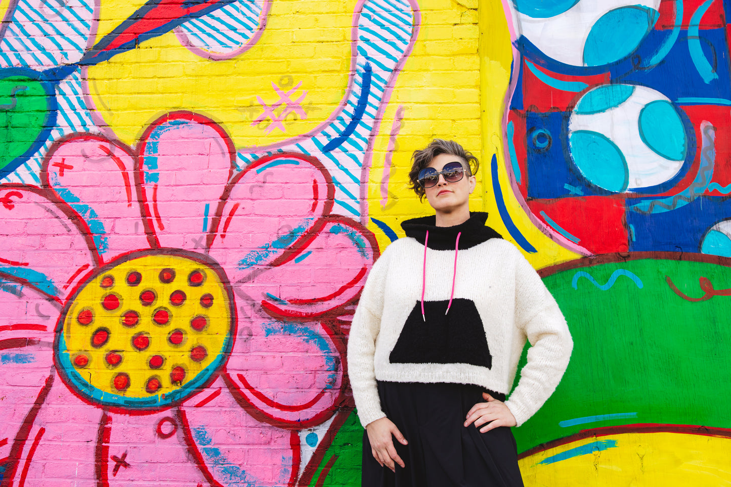 Jen stands in front of a wall mural, one hand on her hip, wearing sunglasses and looking past the camera. She wears an oversized hoodie, knit from black and white yarn.