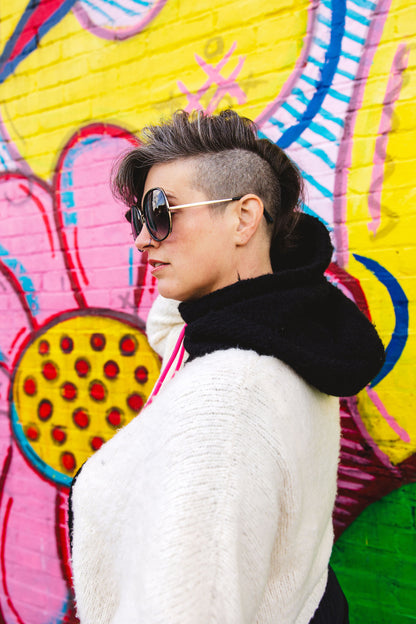 Seen from behind at an angle, Jen wears a soft, hand knit hoodie, made with white yarn and a black hood. A wall mural can be seen in the background.