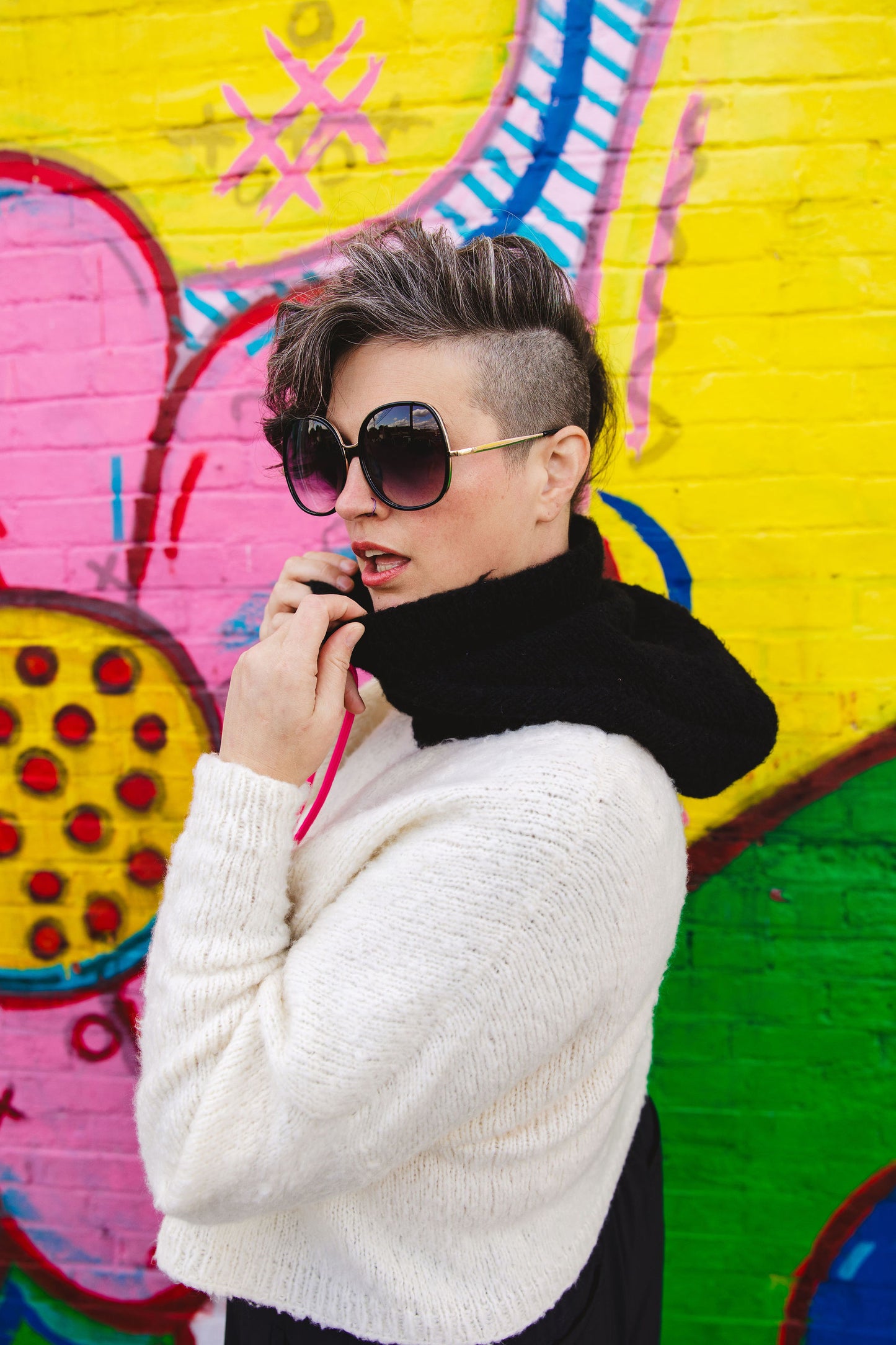 Seen from the side, Jen fingers the soft collar of her black and white knit hoodie. The hoodie is designed in a graphic colorblock effect, featuring neon pink sweater strings. A wall mural can be seen in the background.