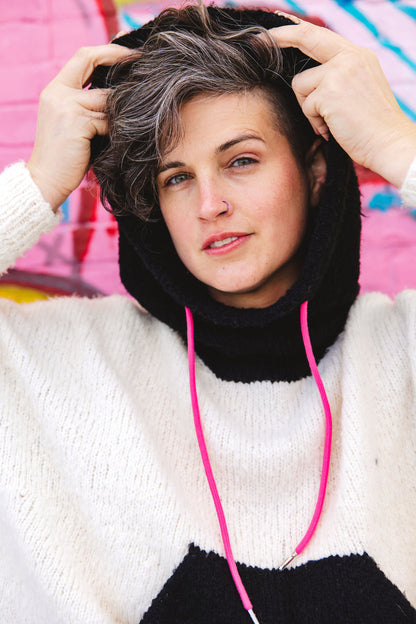 Jen looks at the camera, pulling the hood up on her hand knit sweater. The hood and pouch pocket are knit in black yarn, the body of the sweater knit in white yarn, and features neon pink sweater strings.