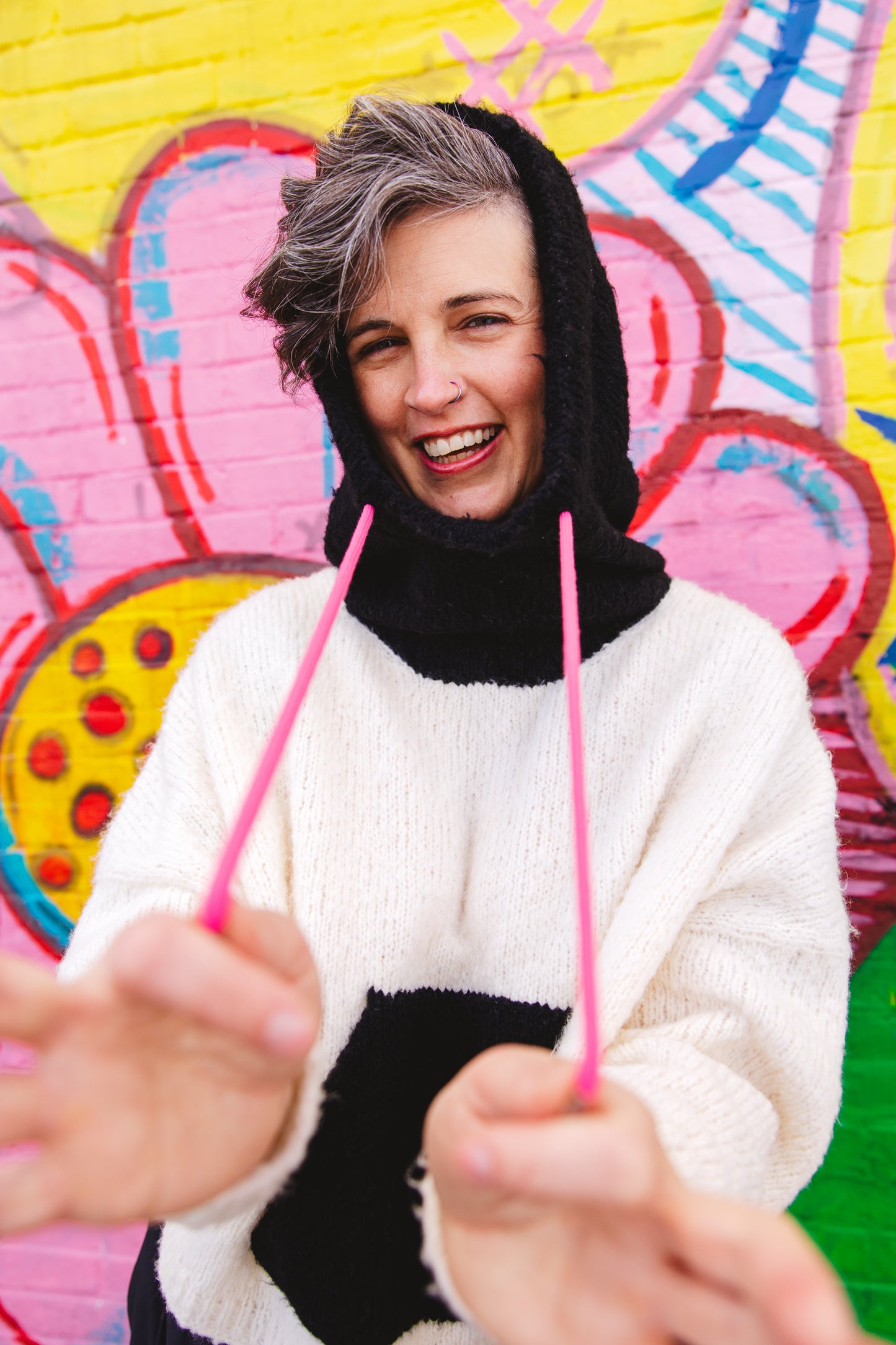 Jen smiles at the camera, pulling on the pink strings of the black and white knit hoodie she wears. A wall mural can be seen in the background.