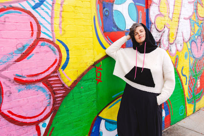 Jen stands, smiling at the camera, wearing the hood up on her black and white hoodie, knit with a pouch pocket at a cropped length. A wall mural can be seen in the background.