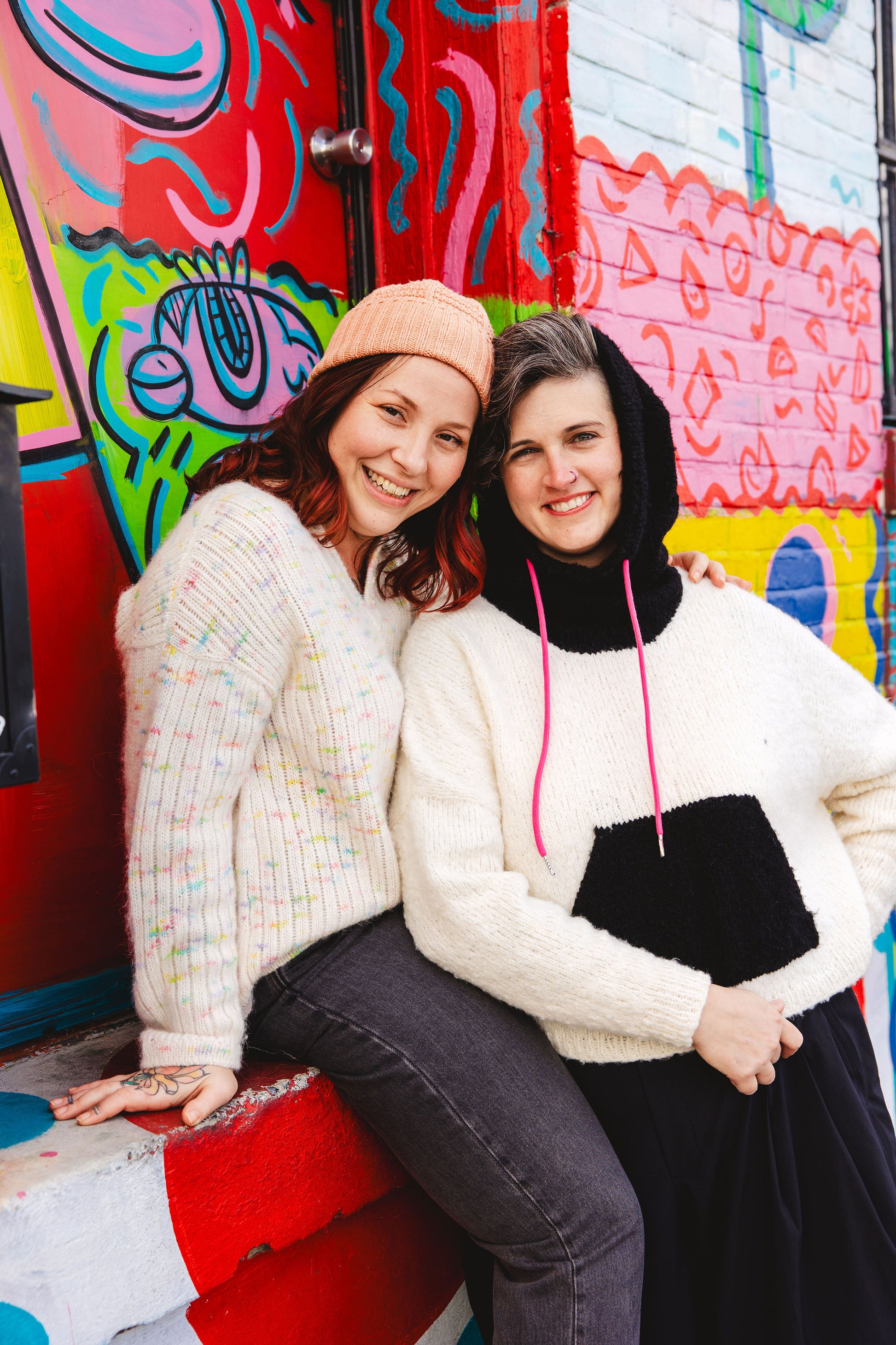 Jen and Bess stand next to an outdoor mural. They are both grinning, and lean together with their heads touching. They both wear soft, fluffy drop shoulder knits.