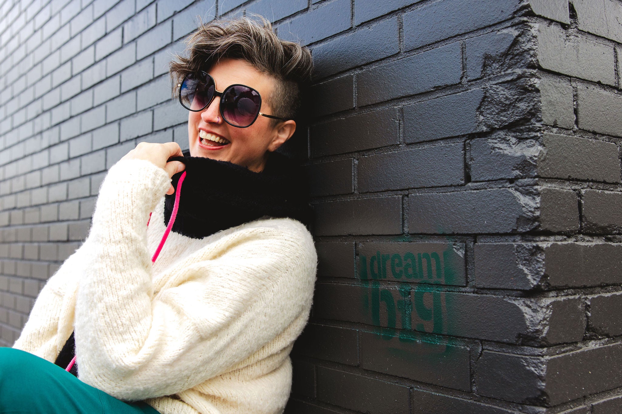 Jen, laughing and leaning against a black brick wall, wears a fluffy knit hoodie. The hoodie is made with white yarn and a contrasting black hood and kangaroo pocket, with neon pink sweater strings.