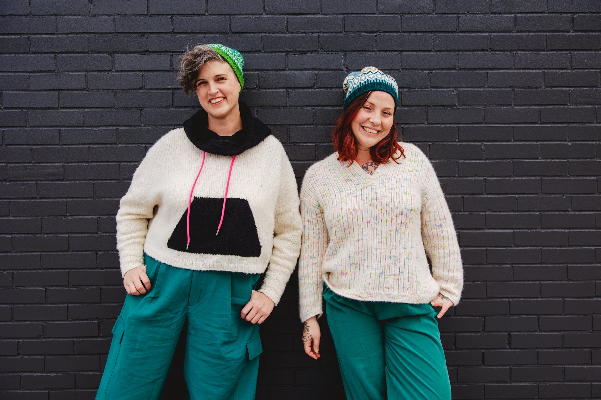 Jen and Bess stand against a brick wall, both grinning at the camera. Jen wears a black and white knit hoodie with a pink hoodie tie, along with a colorwork green and white knit hat. Beside her, Bess wears a white knit henley, made with yarn that has pieces of rainbow tweed, with a blue, orange, and white colorwork knit hat. They both wear matching teal pants.