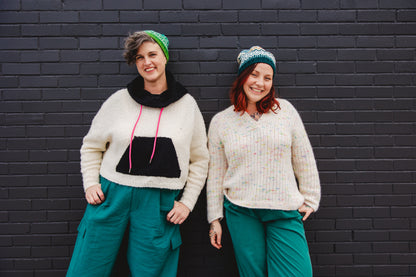 Jen and Bess stand against a brick wall, both grinning at the camera. Jen wears a black and white knit hoodie with a pink hoodie tie, along with a colorwork green and white knit hat. Beside her, Bess wears a white knit henley, made with yarn that has pieces of rainbow tweed, with a blue, orange, and white colorwork knit hat. They both wear matching teal pants.