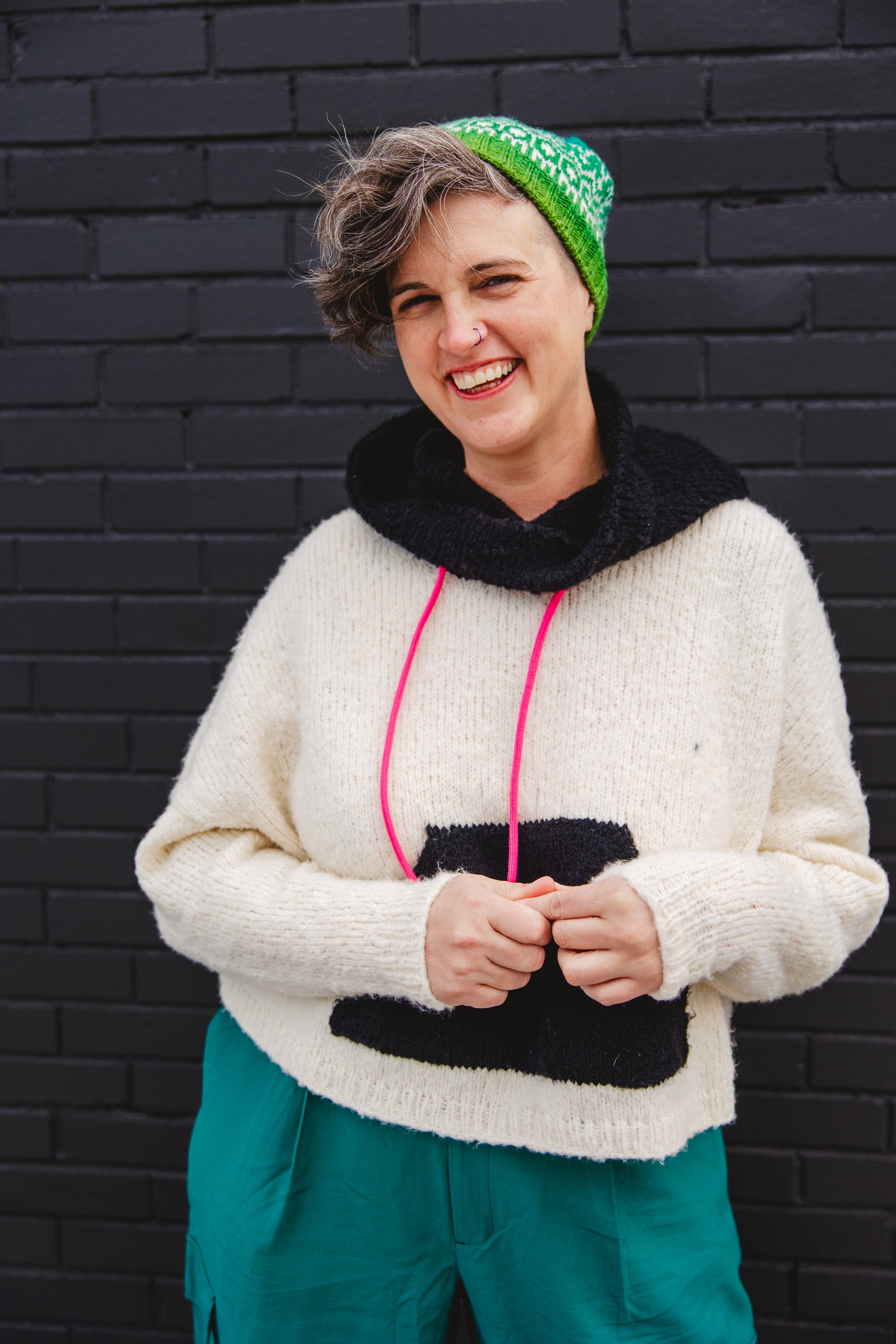Jen stands, laughing, in front of a grey brick wall. She wears a white hoodie, knit with a black hood and pouch, that has pink hoodie strings. Her pants are teal, and her hat is a green and white colorwork beanie.