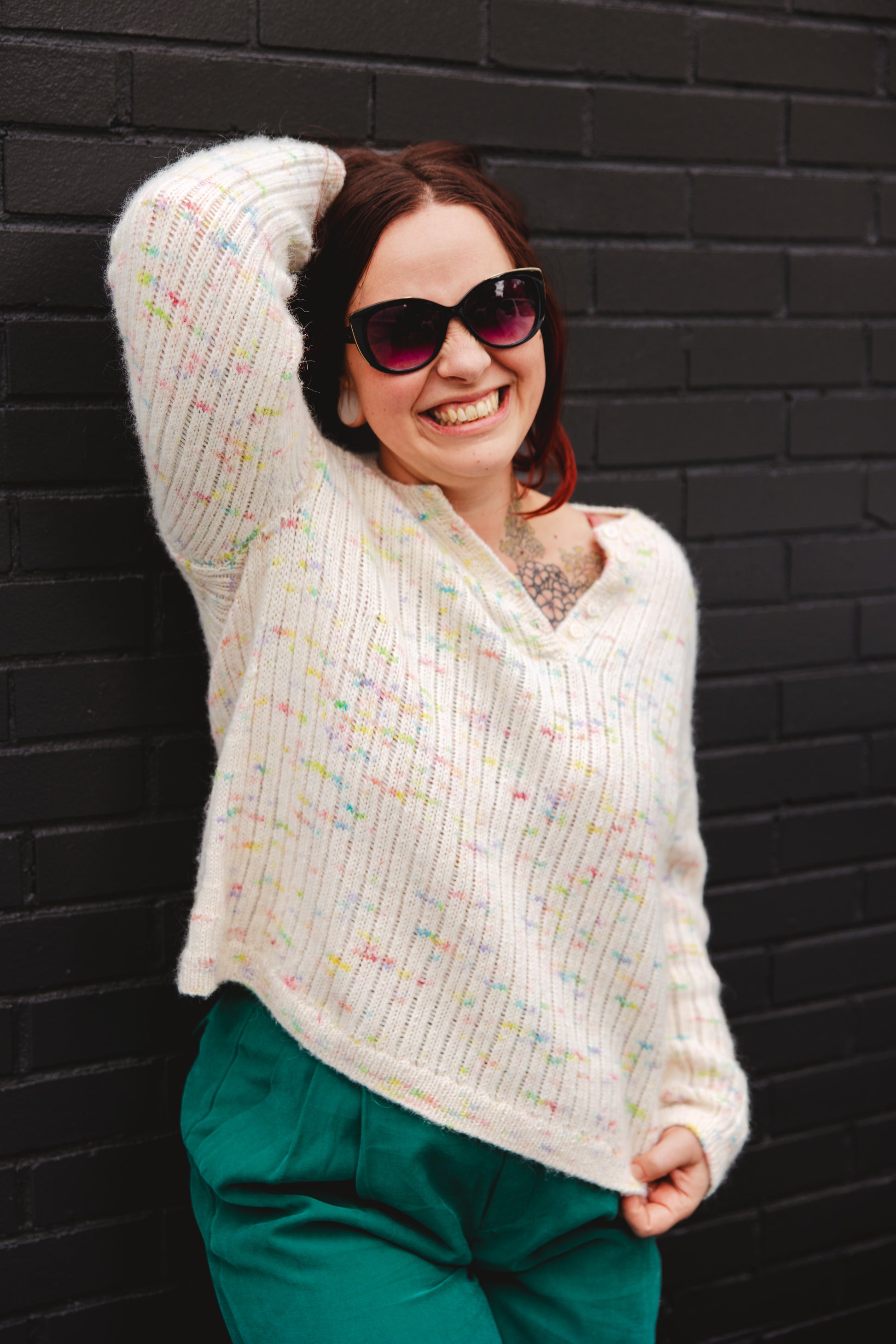 Bess grins at the camera, one hand in her hair. Her henley shirt is knit with a white yarn with pieces of rainbow tweed, and her pants are teal.