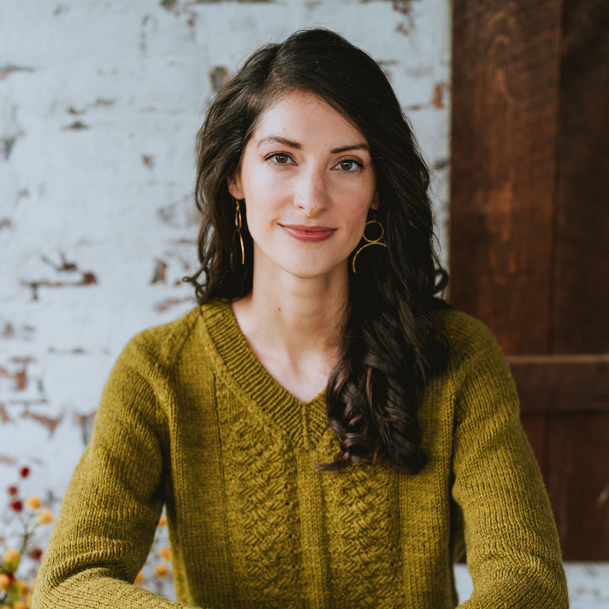 Laura sits, smiling at the camera, wearing a golden colored hand knit sweater. The sweater has two cable braids down the front and a ribbed V-neck neckband.