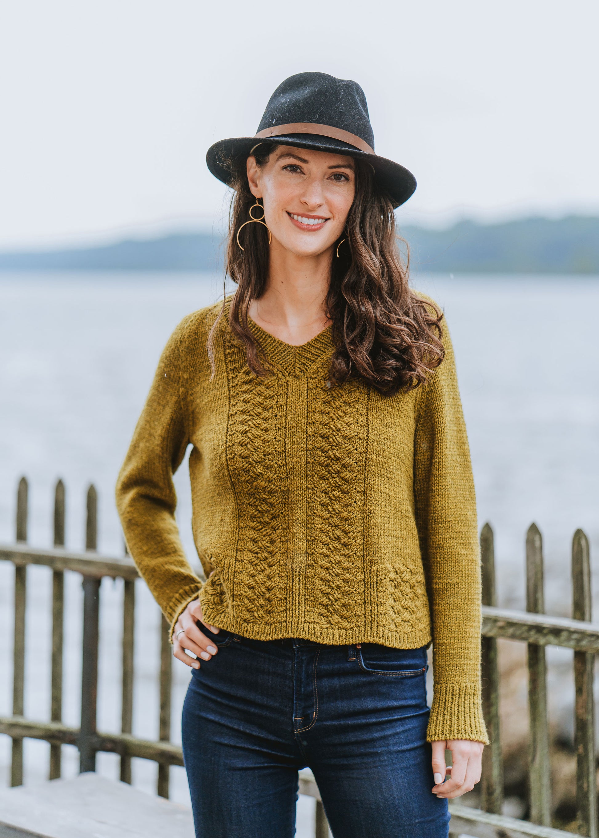 Laura, a woman with long brunette hair, stands in front of a short fence, a body of water and hills in the background. She wears a hand knit sweater featuring two cable strips down the front and a V-neck.
