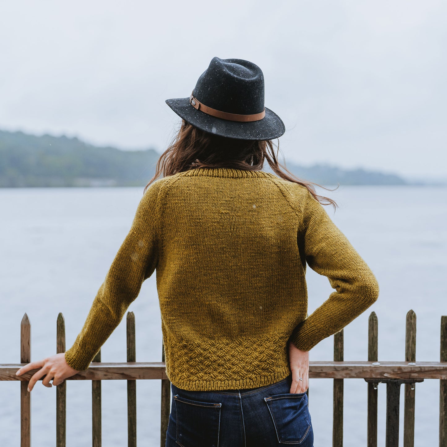 Seen from behind, Laura stands in front of a body of water. The camera focuses on the details of her knit sweater, showing off the cable plait design around the hem. 