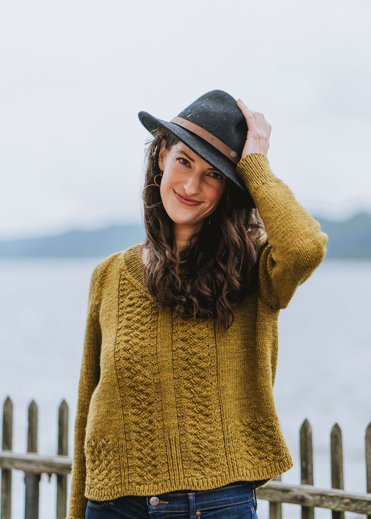 Laura smiles at the camera, one hand on her hat. Her golden hand knit sweater features cable details down the front and around the hem. 