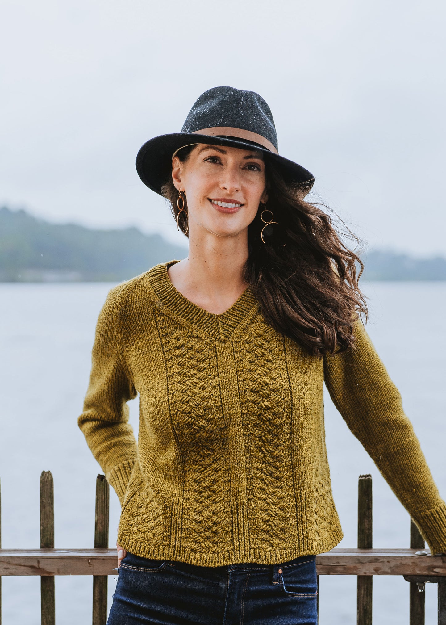 Laura stands outside on front of a body of water, smiling at the camera. Her handmade sweater is knit up with golden colored yarn, with cable plait details down the front.