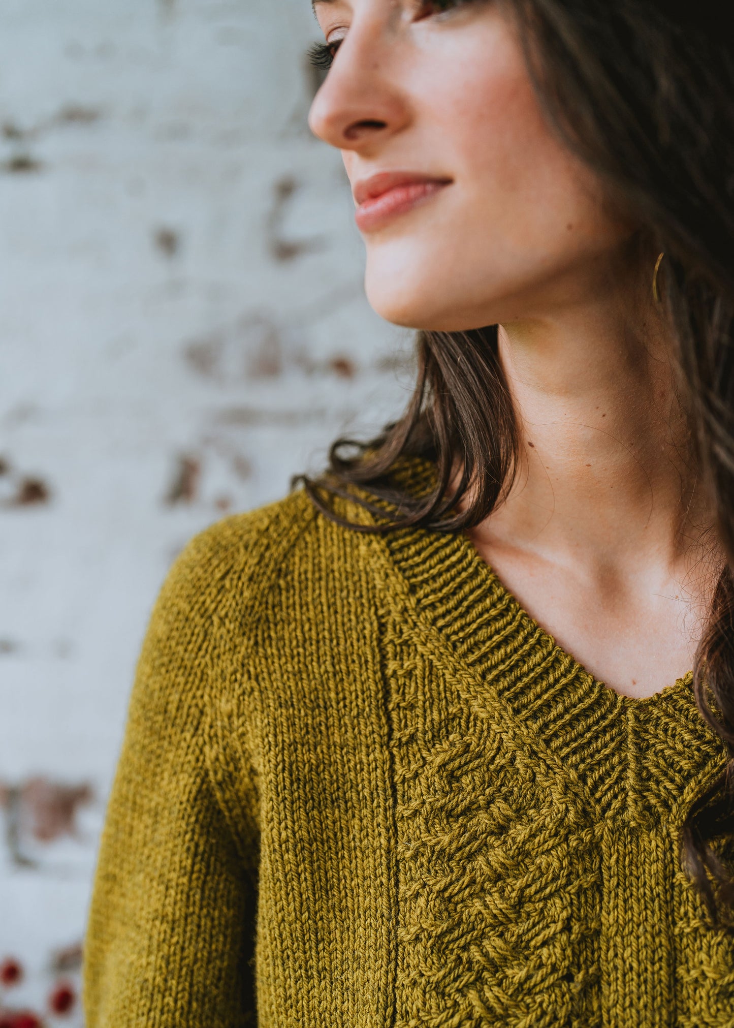 Seen from close up, Laura looks off camera, wearing a golden colored hand knit sweater. The sweater is knit up with cable details and a V-neck.