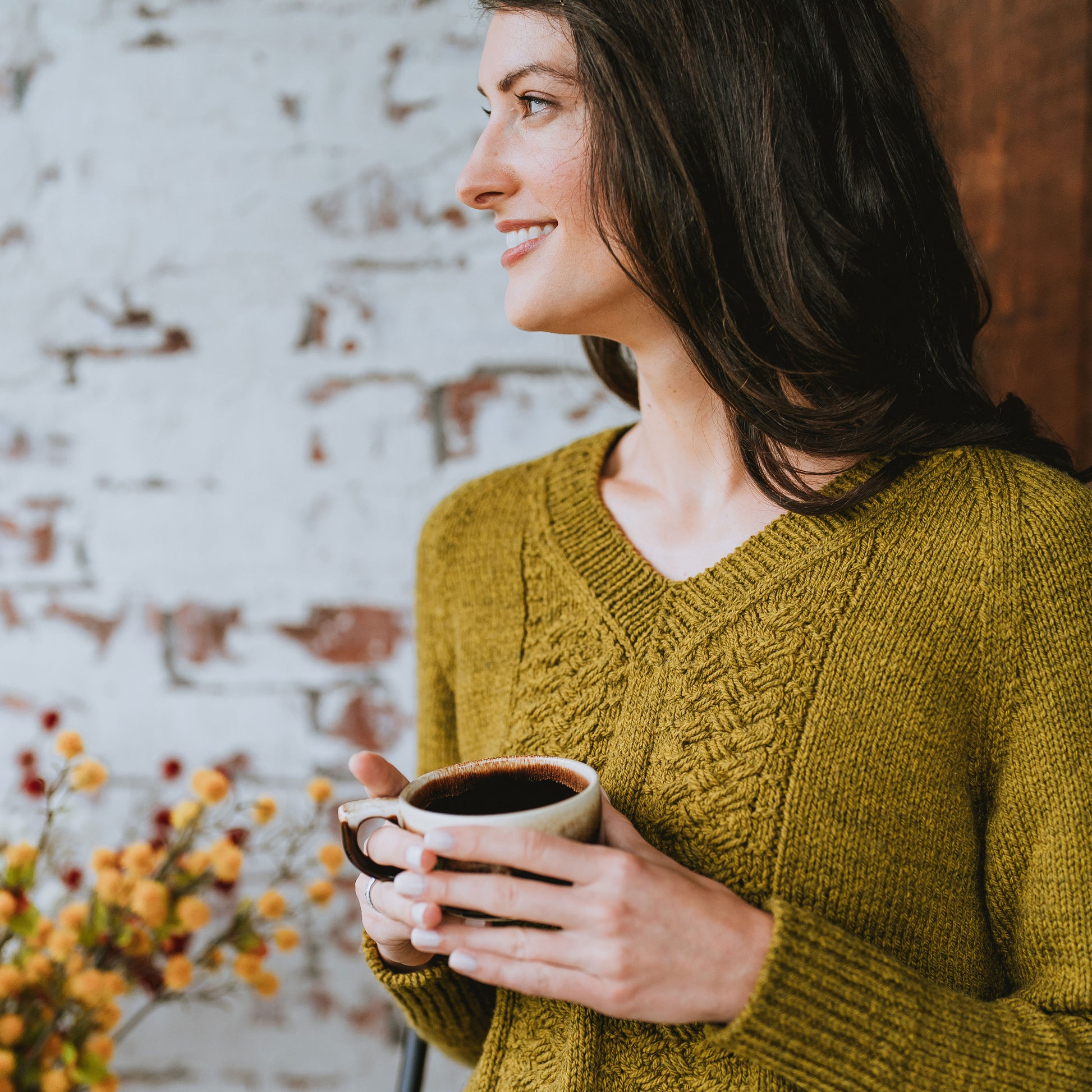Seen at a three quarter angle, Laura looks off camera, holding a cup of coffee. She wears a hand knit sweater with cable details and a V-neck.