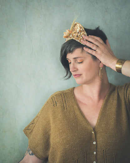 Jen, standing in front of a grey wall, wears a dolman style tee, knit with lace strips around the shoulders and hems and a lace panel down the sleeves. The yarn is a light gold color.