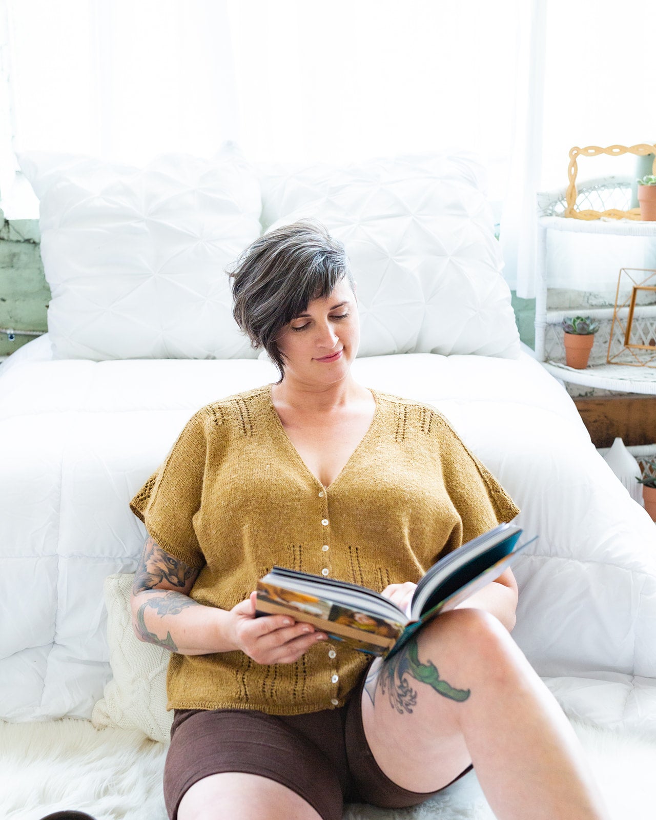 Jen sits at the end of a chaise lounge, reading a book. She wears black shorts with a gold tee, buttoned with small pearl buttons and adorned with eyelet lace strips coming down from the shoulders and up from the hem.
