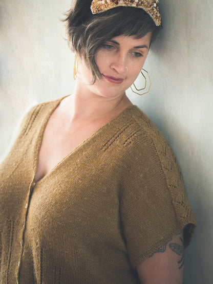 Jen leans against a wall, wearing a gold button tee, designed in a dolman style. The sleeves feature a lacy design, and eyelet lace strips coming down from the shoulders and up from the hem.