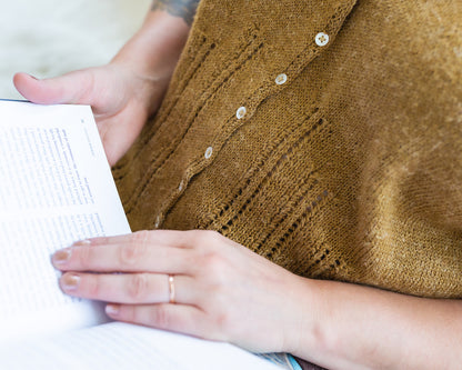 Jen, seen from the side, reads a book. She wears a dolman tee, closed with small pearl buttons and knit from gold yarn. Eyelact lace strips rise from the hem.