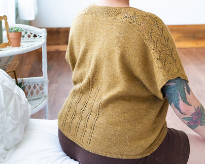 Jen sits on a chaise, seen from behind. She wears black shorts and a gold dolman tee. The tee has lace strips down the sleeves and up towards the middle of the back. Eyelet lace strips rise up from the thin ribbed hem.