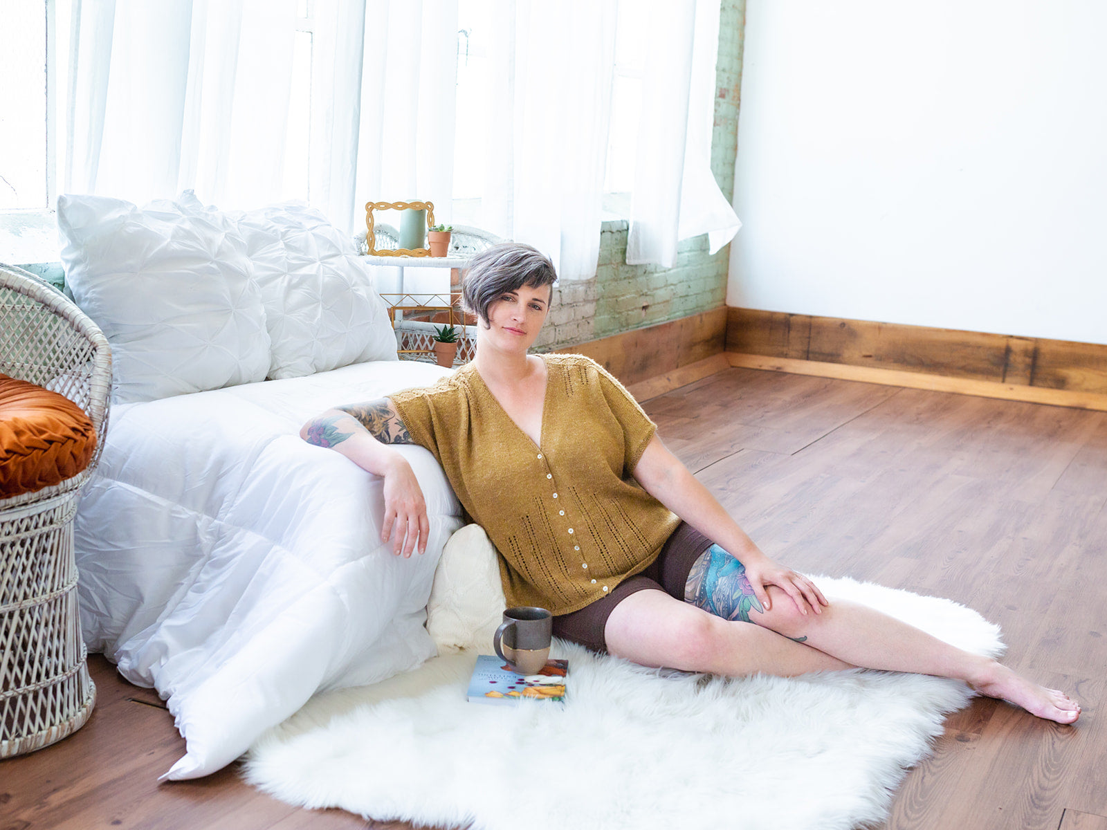 Jen seats on a sheepskin, leaning against the end of a chaise. She smiles at the camera, wearing black shorts with a gold, hand knit tee. The tee is designed in a dolman style, buttoned with small pearl buttons and featuring a lace stitch.