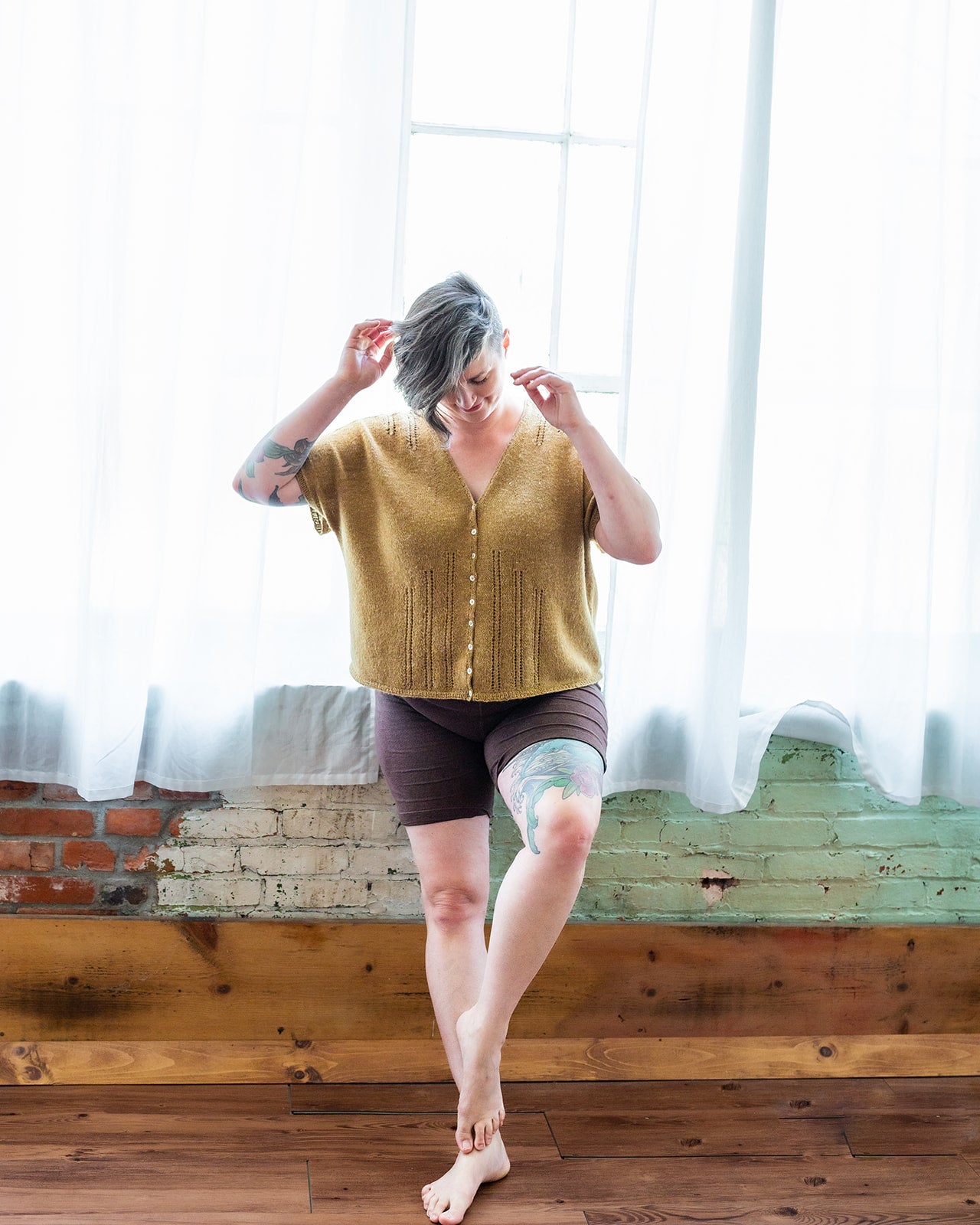 Jen leans against a window, adjusting her hair. She wears black shorts with a gold, dolman style tee. The tee features eyelet lace strips, a button closure, and a V-neckline.