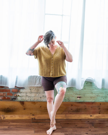 Jen leans against a window, adjusting her hair. She wears black shorts with a gold, dolman style tee. The tee features eyelet lace strips, a button closure, and a V-neckline.