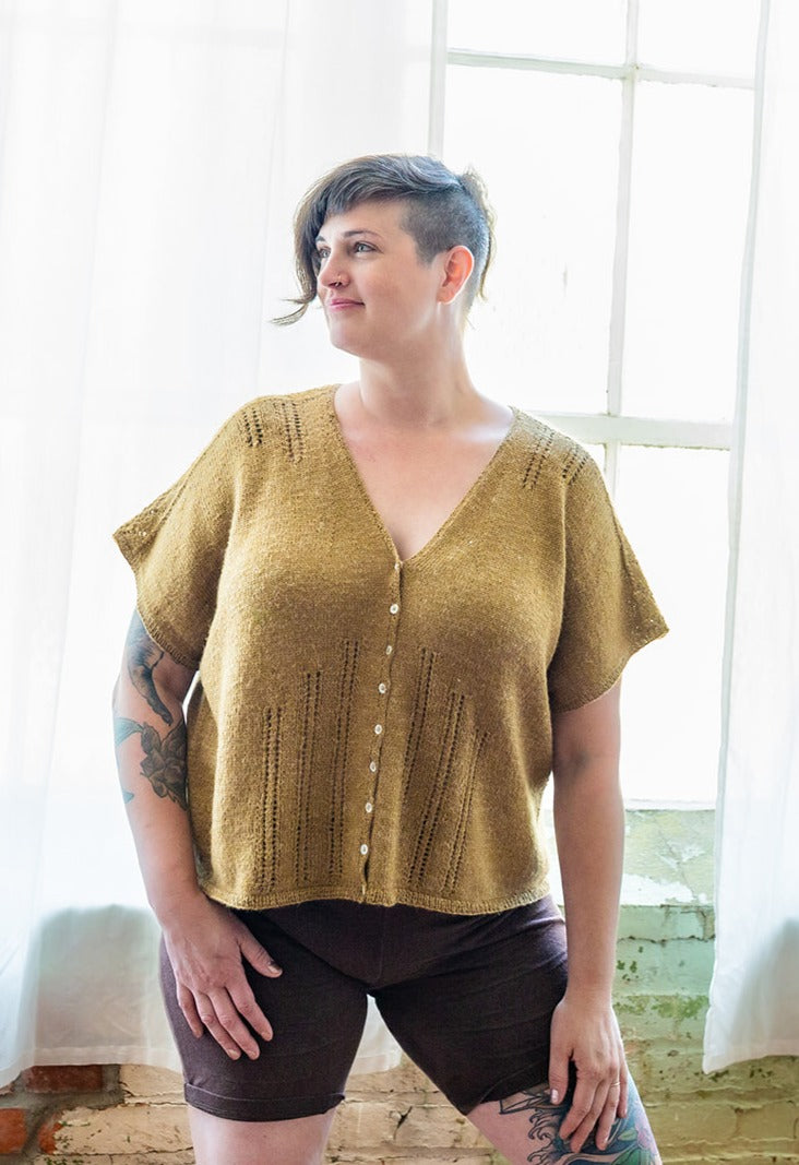 Jen stands, smiling and look off camera, in front of a window. She wears a dolman style tee, knit in gold yarn, with a pair of black shorts. The tee has eyelet lace strips coming down from the shoulders and up from the hem, and is closed with small pearl buttons.