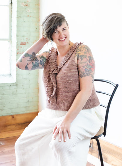 Seated, Jen smiles at the camera, wearing a pink tank top that has a ruffled collar, along with a pair of white flared pants.