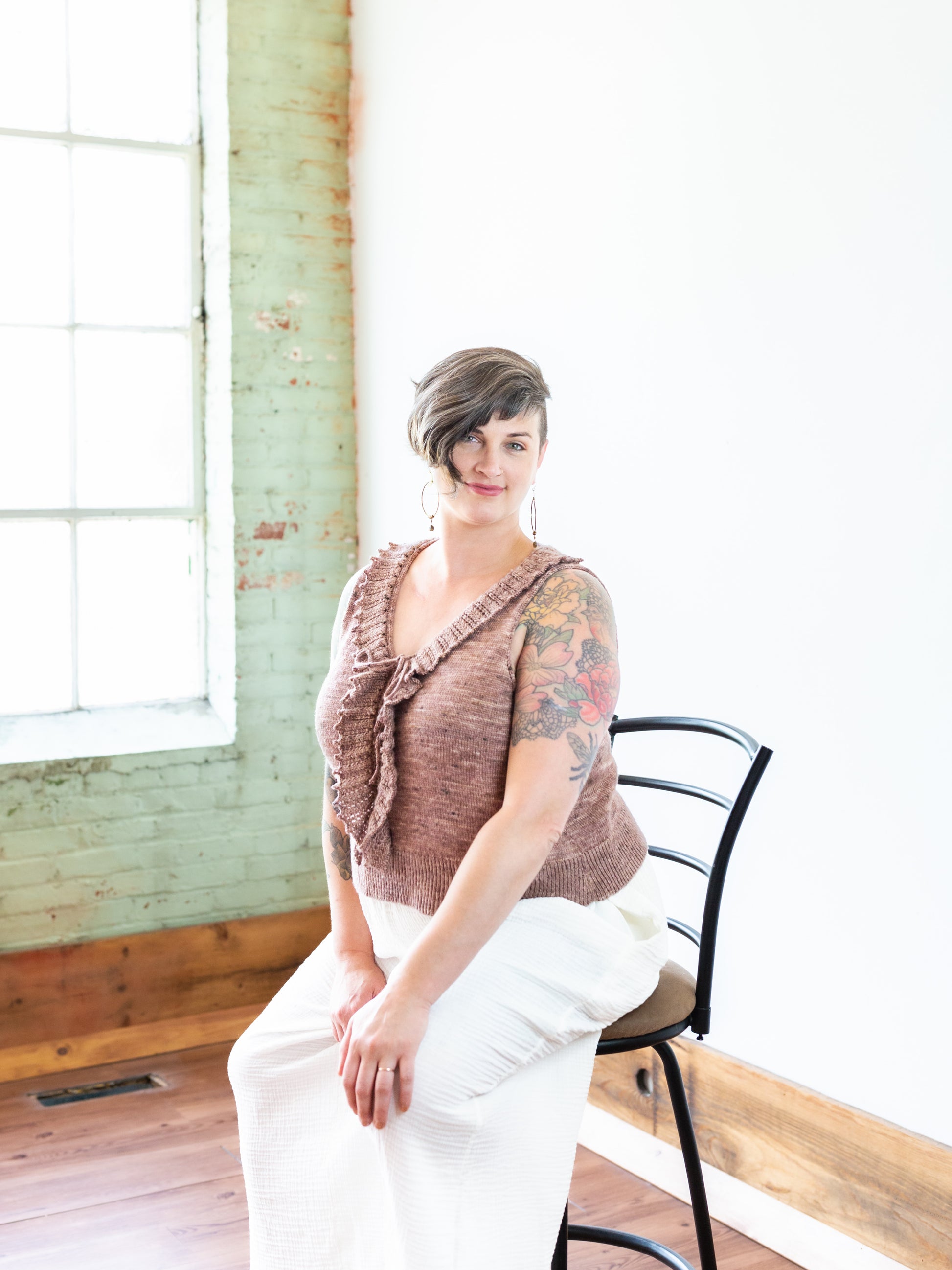 Jen sits on a chair, smiling at a camera, in a pair of white pants and a pink, hand knit tank top. The top has a ruffled lace collar.