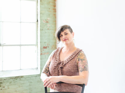 Jen leans on a chair, smiling at the camera. She wears a dusty pink tank top, knit with a ruffle collar and a bow tied in the front.