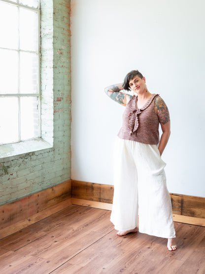 Jen stands in a room with wood floors, smiling at the camera. She wears a silky, ruffled, hand knit top with white flared pants.