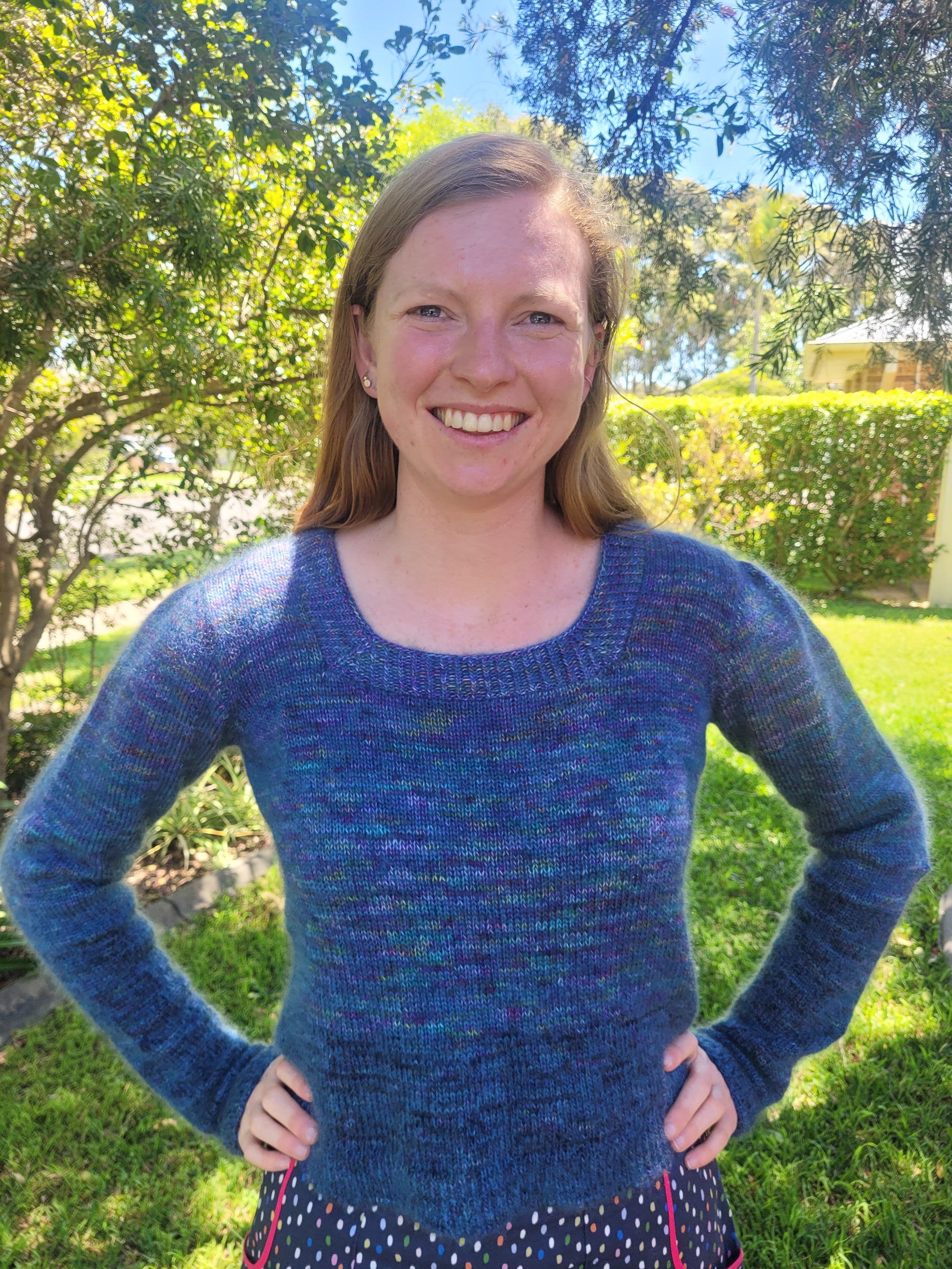 Ellen stands outside, smiling at the camera. She wears a blue sweater, knit with a square neck and gathered sleeves. The yarn has a slight halo to it, which shines in the sun.