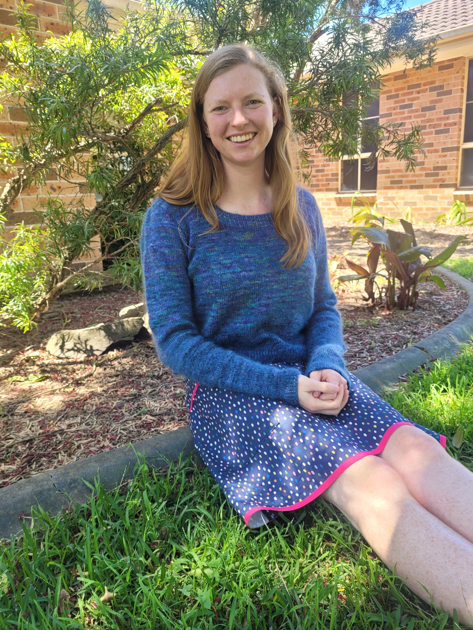 Ellen sits outside near a garden, smiling at the camera. She wears a blue skirt with a hand knit sweater. The sweater is made with blue yarn, and has sleeves that are gathered at the top seams.
