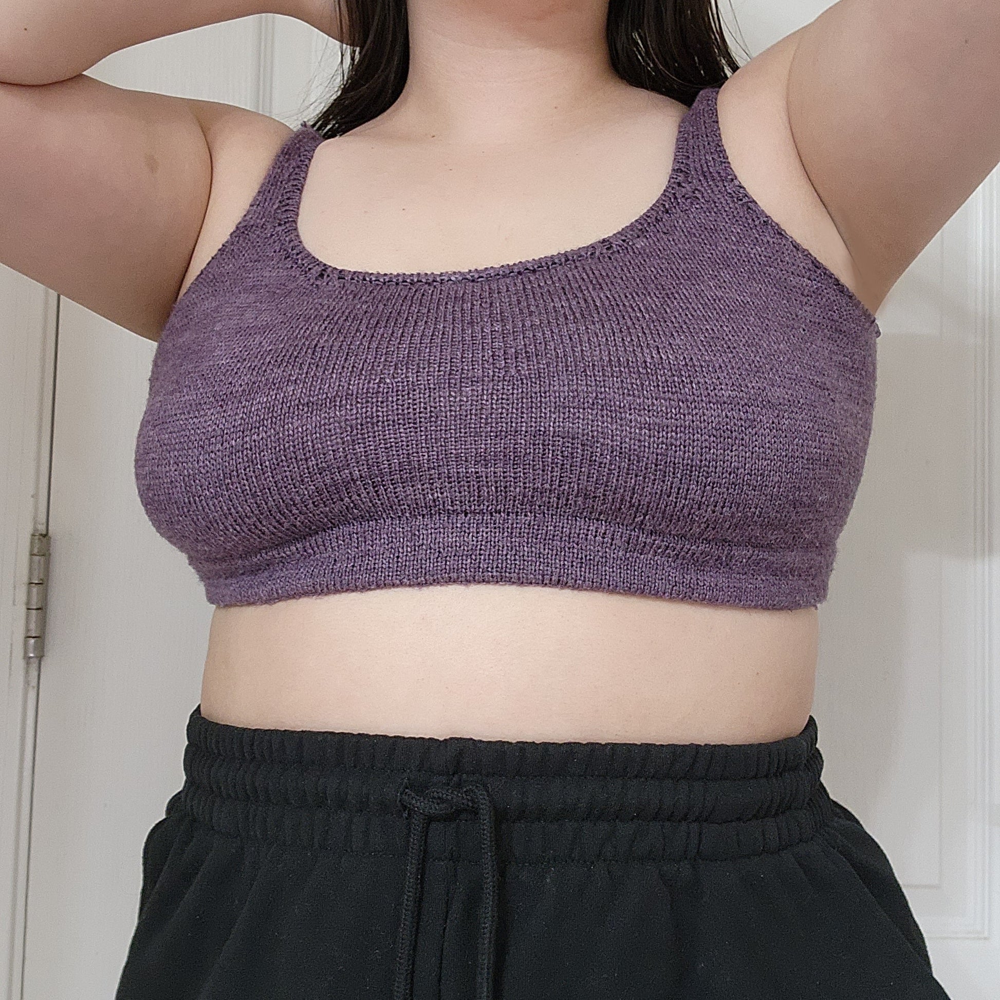 Jordynn, wearing a deep purple, hand knit bralette, is seen from the shoulders down. She has both hands in her black hair, and wears a pair of black sweatpants.
