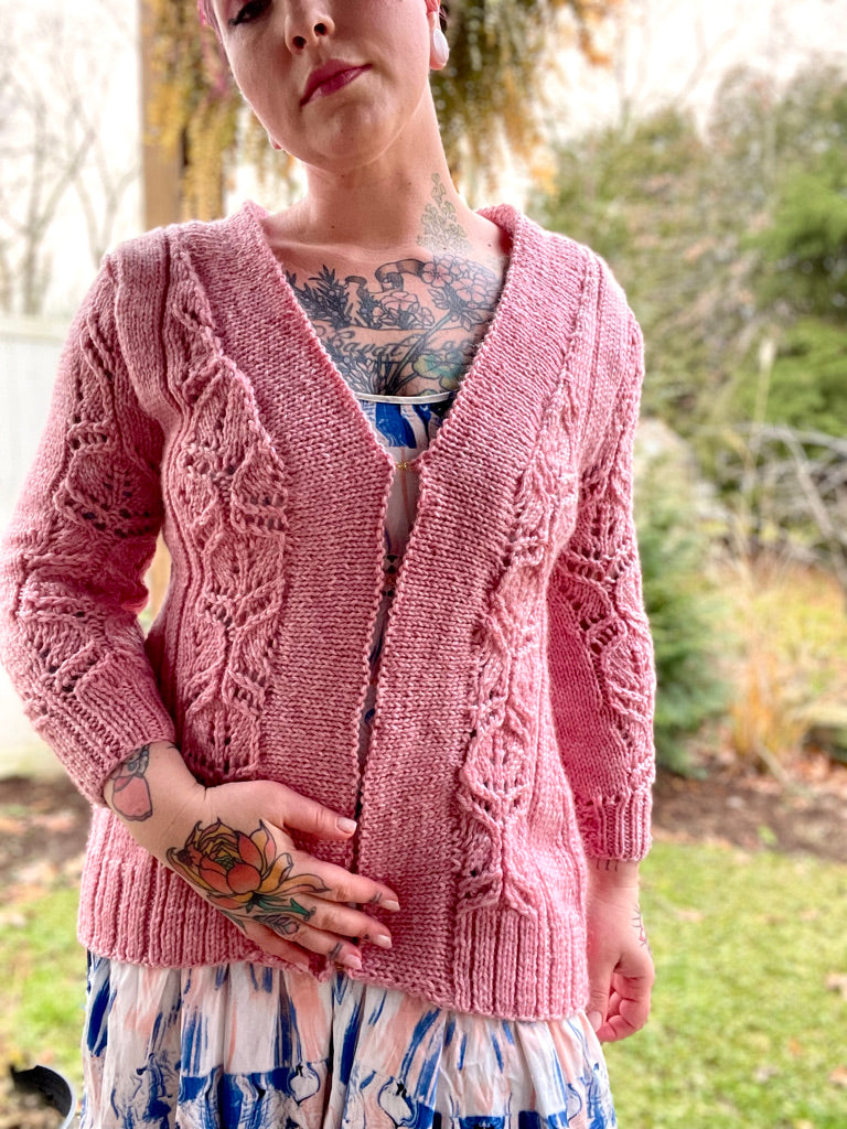 Bess wears the Katherine cardigan, a light pink sweater knit in lace panels with bulky yarn. A backyard can be seen in the background.