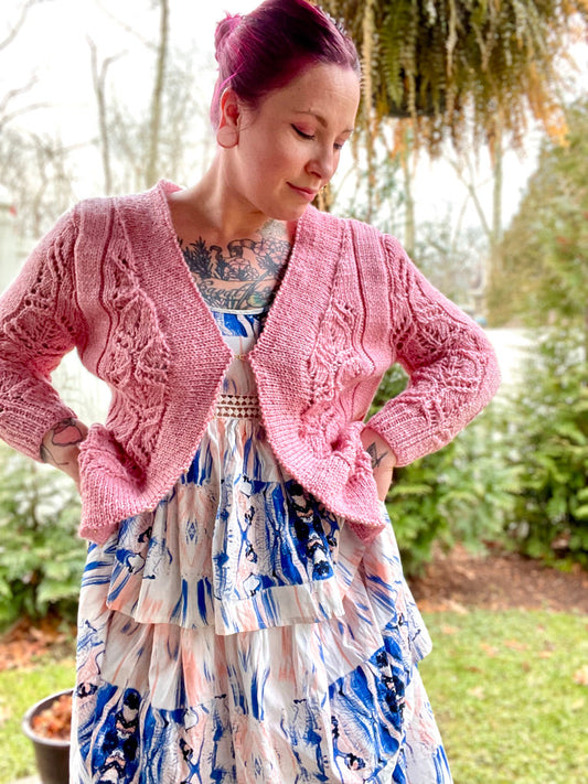 Bess wears a light pink, bulky knit cardigan over a white and blue dress. The cardigan features lace panels and is held together with a simple front closure.