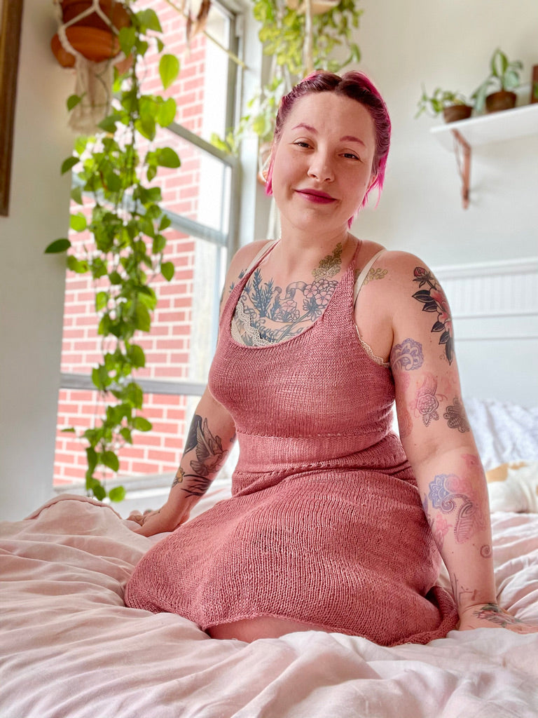 Bess sits on a light pink comforter, houseplants and a window in the background. She wears a hand knit, dusty pink sleeveless dress, and smiles at the camera.