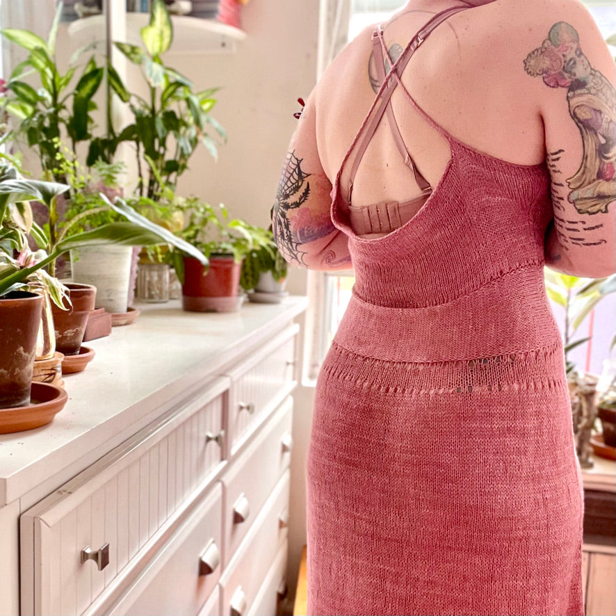Bess stands with her back to the camera, showing off the scooped back and crossed straps of her hand knit dress. The dress has a cinched waistband and is knit from silky yarn. A dresser with houseplants on it can be seen in the background.
