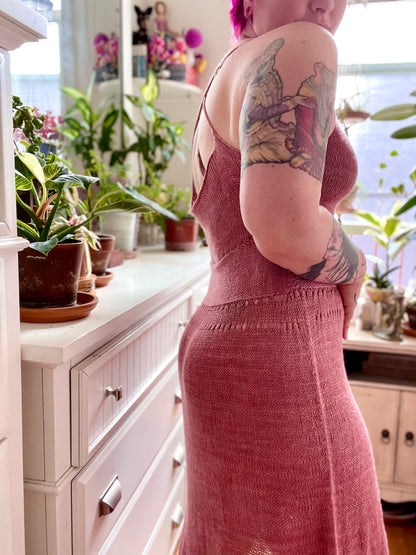 Seen from the shoulders down, Bess stands at an angle to camera. She wears a dusty pink sleeves dress, the straps crossing in the back. The dress is hand knit with a sculpted waist.