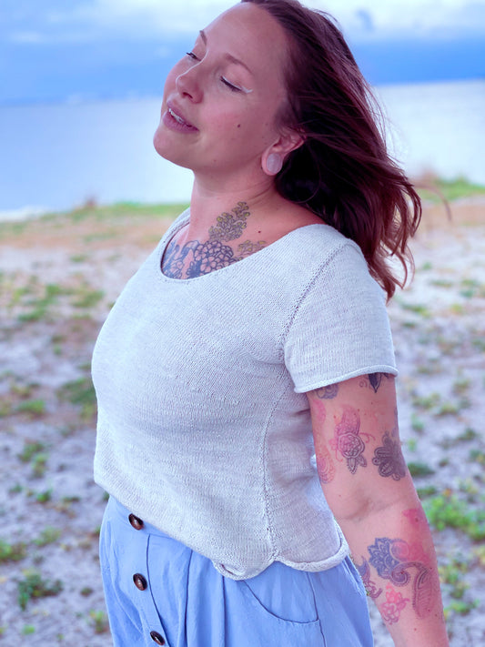 Bess stands on a shoreline at a three quarter angle to the camera. She wears a white knit tee with a blue skirt.