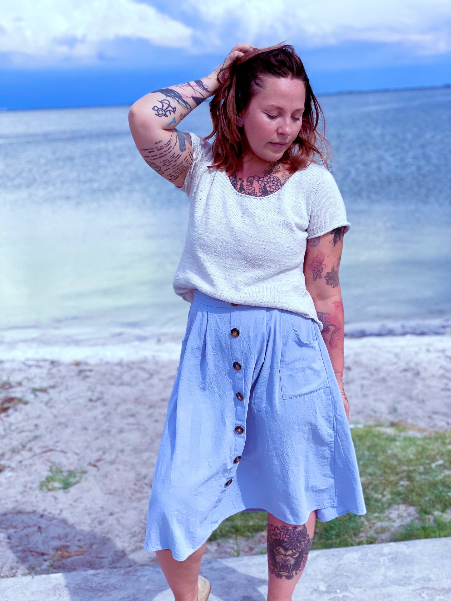 Bess stands, a body of water behind her, wearing a white knit tee with a light blue midi skirt.