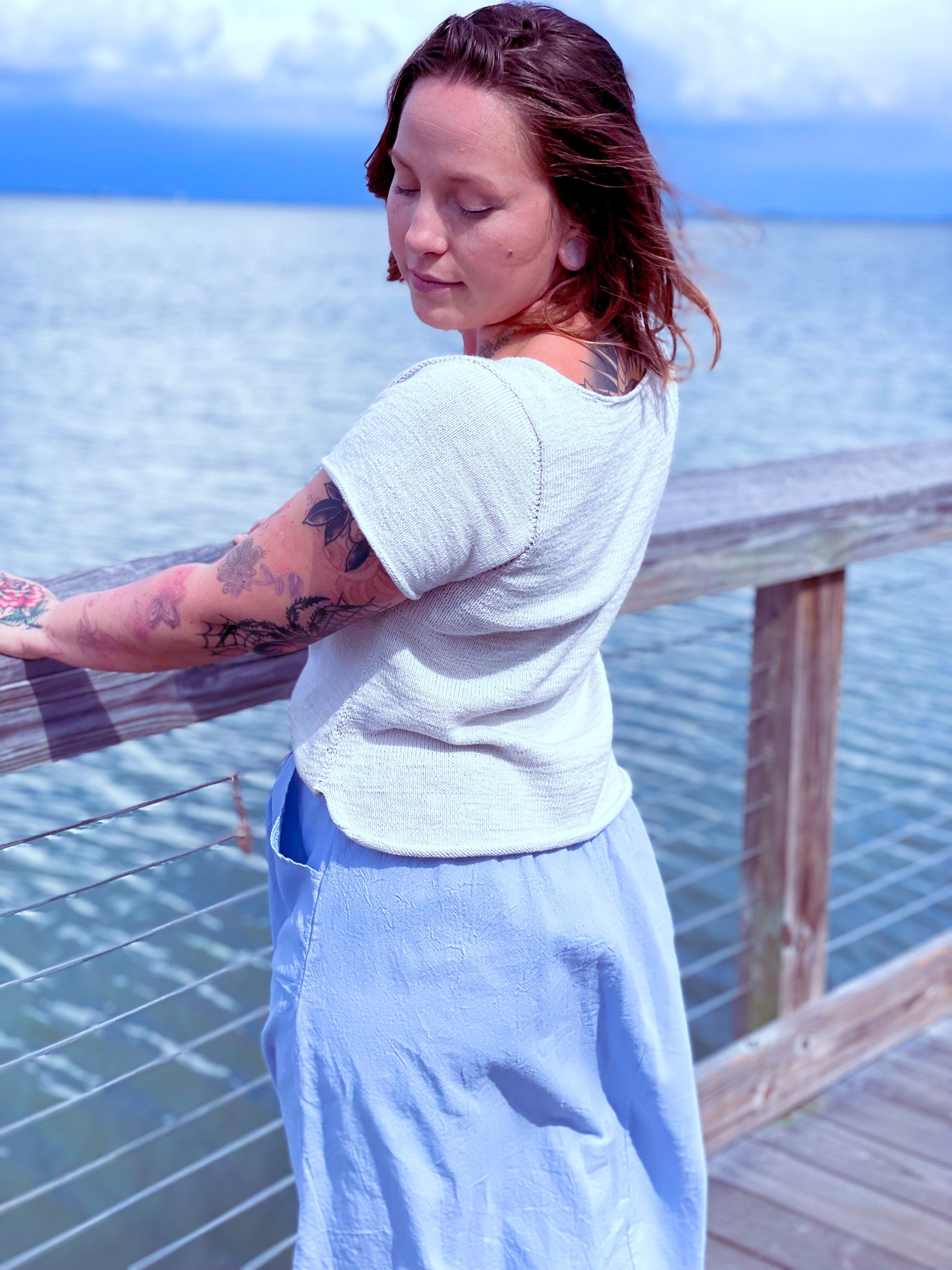 Bess stands on a bridge, her back to the camera. She wears a white, hand knit tee with a light blue skirt.