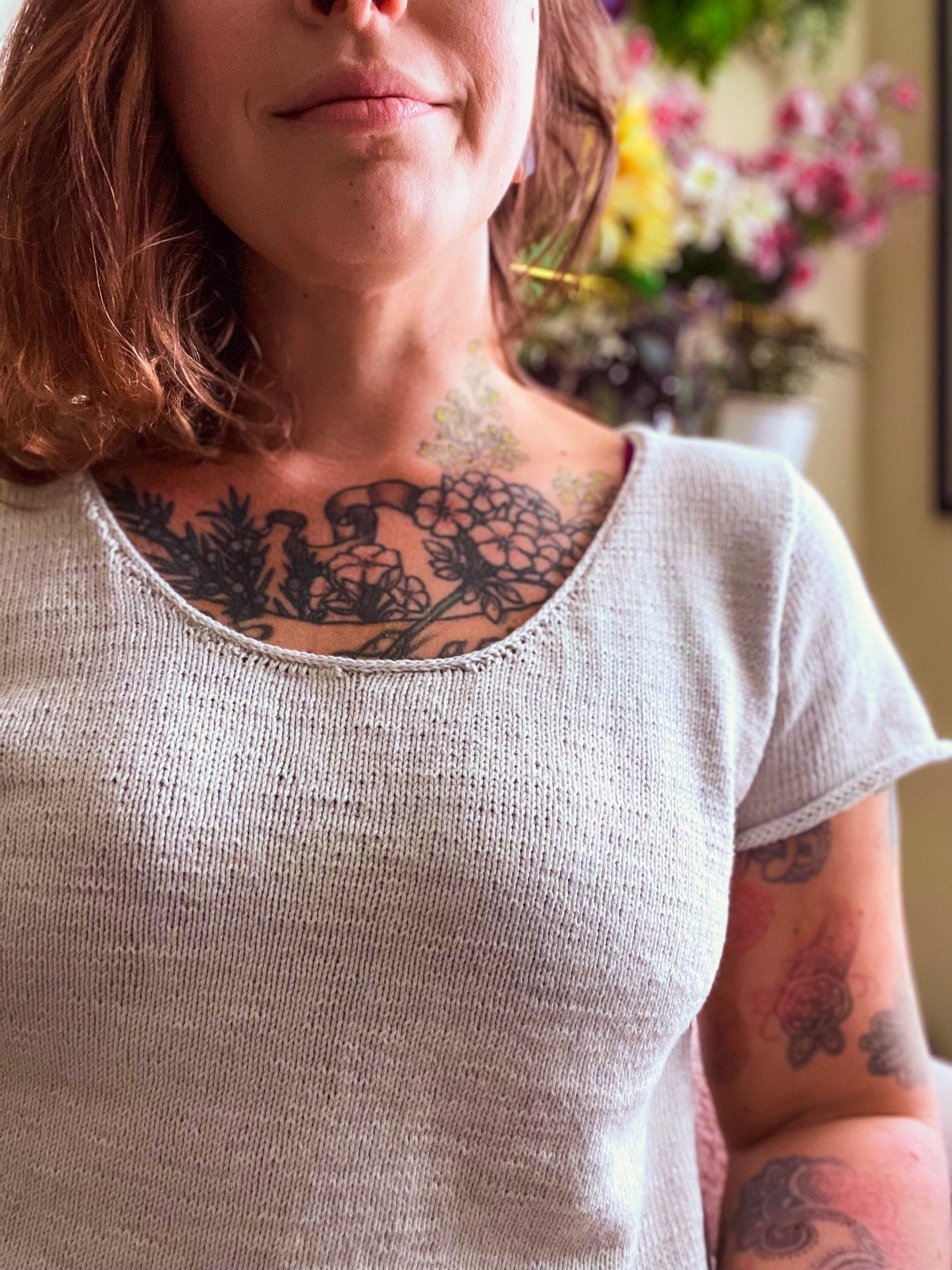 Seen close up, Bess wears a white, hand knit tee with an i-cord egde on the neckline and sleeves. Flowers can be seen in the background.