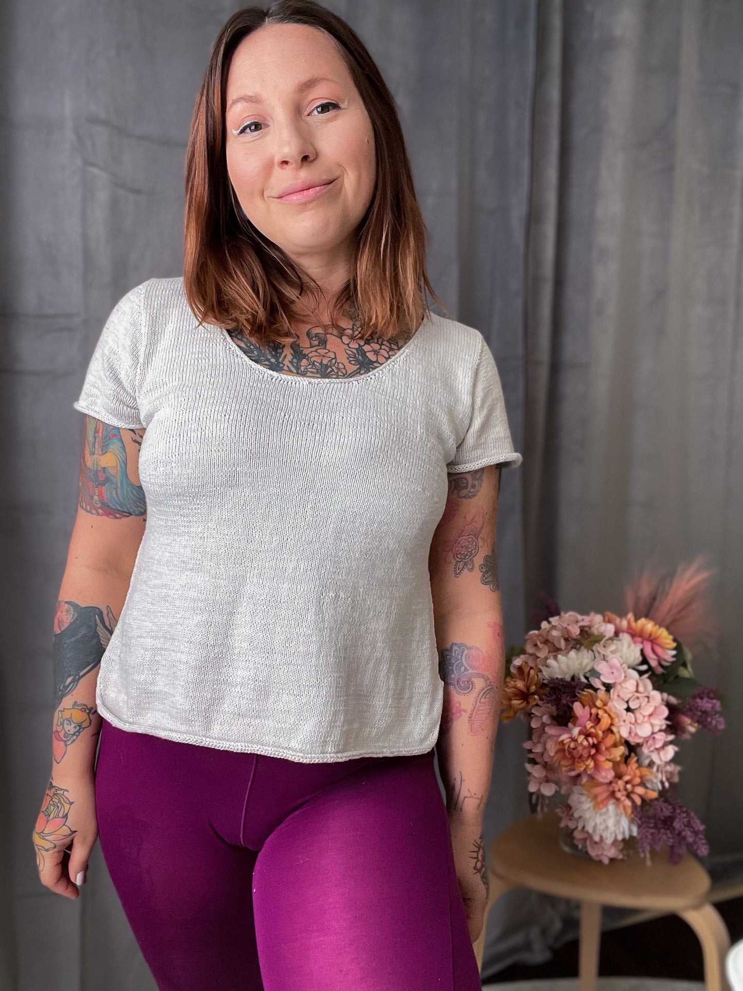 Bess smiles at the camera, wearing a white knit tee with magenta leggings. Flowers on a coffee table can be seen in the background.