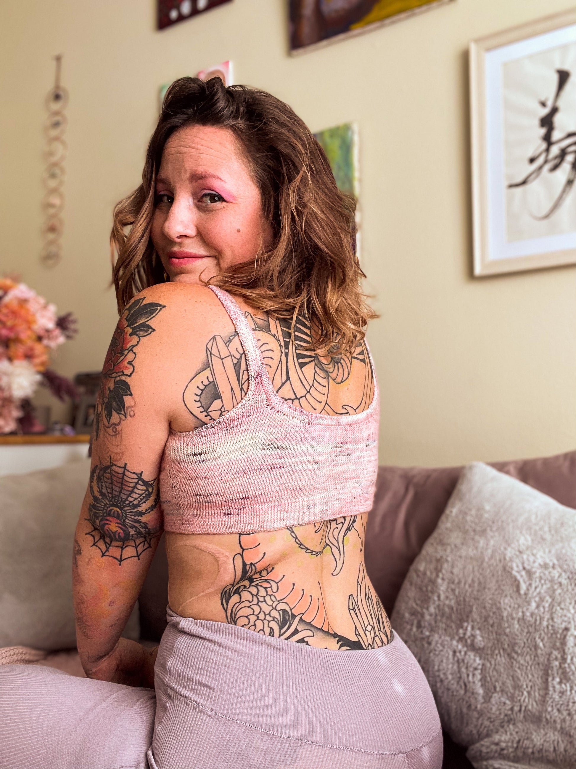 Seen from behind at an angle, Bess wears a hand knit, light pink speckled bralette with matching leggings. She turns her head to smile at the camera.