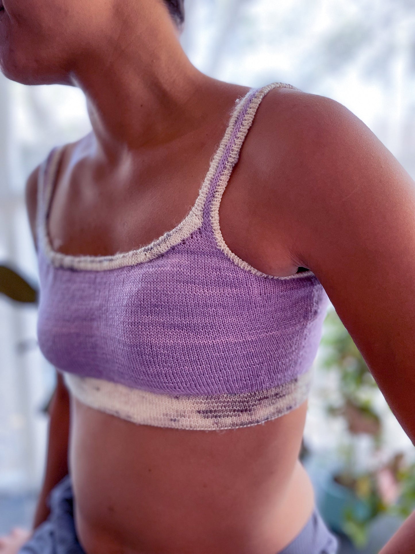Seen close up, Candace wears a light purple bralette, knit with a contrasting speckled white band and straps. Houseplants can be seen in the background.